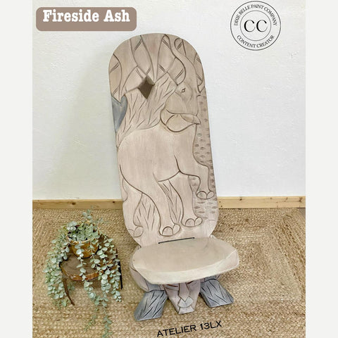 A uniquely designed chair with an intricately carved elephant and foliage on the backrest, stained in a pale color. The seat is surrounded by greenery on a woven mat. The top left corner displays the text "Fireside Ash."
