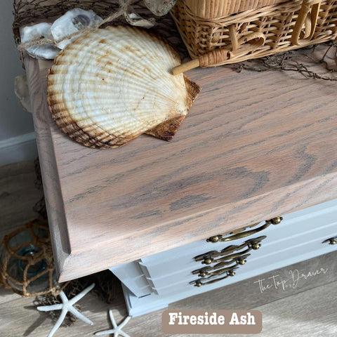 A close-up of a light wood cabinet displaying coastal decor. Sea shells, a fishing net, and a wicker basket adorn the top. A label reads "Fireside Ash" near the wood. Nautical elements like a starfish and rope are visible.
