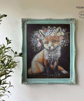 A decorative painting of a fox with a crown of colorful flowers is displayed in an ornate turquoise frame. The artwork is mounted on a white wall, with a leafy green plant placed to the left of the painting.