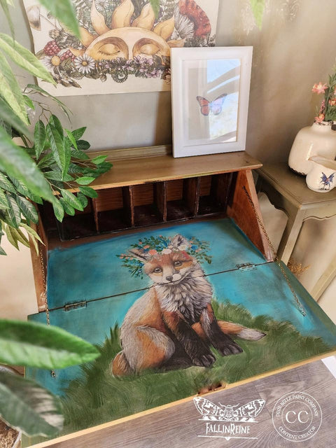 A wooden desk with an open compartment reveals a painted image of a fox with flowers on its head, set against a blue background. On top of the desk are framed butterfly art and a potted plant. The wall behind it has sun and floral decorations.