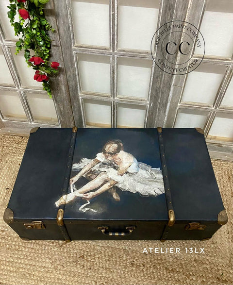 A suitcase with a painting of a seated ballerina adjusting her ballet slippers on the lid. The chest is placed on a woven rug in front of a decorative window pane with red flowers. The logos "CC/ Dixie Belle Paint Company Content Creator" and "Atelier 13LX" are visible near the chest.