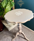 A small, round, light beige vintage side table with scalloped edges and white floral detailing on top stands on a three-legged pedestal base next to a green leafy plant on a carpeted floor.