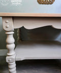 A close-up of a light beige wooden table with ornate, turned legs and a scalloped apron. The table sits on a dark wood floor, and logos are visible in the top left corner of the image.
