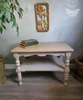 A painted wooden table with ornate legs stands on a gray floor. Three stacked vintage books sit on top. A decorative framed artwork hangs on the wall above, near potted plants.