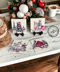 A table displays festive holiday coasters with the Frosty Forest Friends Decoupage Paper. Each coaster features a winter scene: a dog in a red Santa hat, a bear in a red scarf and hat next to gifts, a moose in a snowy field, and a red cabin with a family of deer in front. A centerpiece with ornaments, a cup of coffee, and rustic decor accents the setting.