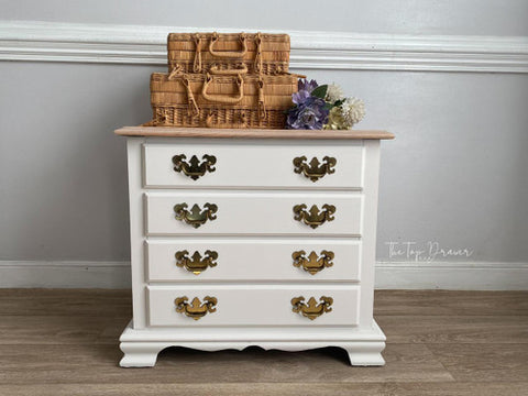 A small white dresser with four drawers featuring ornate brass handles stands against a gray wall. On top, there are three woven baskets and a bouquet of purple and white flowers. The floor is wooden.