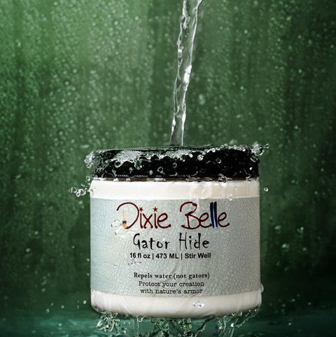 A jar of Dixie Belle Gator Hide is splashed with a stream of water against a green background, demonstrating its water-repellent properties. Water droplets are seen around and on the container.