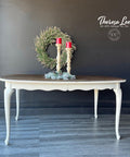 Grayish white painted coffee table with a medium wood top. Staged with candlesticks and garland on top. A green wreath hangs behind the piece on the wall.