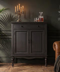 Moody vintage-inspired room featuring a dark painted cabinet with candlelit decor, crystal decanter set, leather chair, globe, and lush greenery against deep charcoal walls and warm wood flooring.