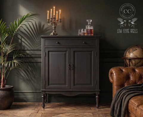 Moody vintage-inspired room featuring a dark painted cabinet with candlelit decor, crystal decanter set, leather chair, globe, and lush greenery against deep charcoal walls and warm wood flooring.