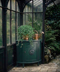 Dark blue-green vintage cabinet styled with a cascading plant and copper watering can inside a moody greenhouse filled with lush greenery and rain-kissed glass windows.