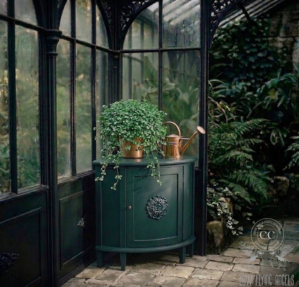 Dark blue-green vintage cabinet styled with a cascading plant and copper watering can inside a moody greenhouse filled with lush greenery and rain-kissed glass windows.