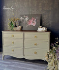 A light olive six-drawer dresser with gold knobs stands against a dark, patterned wall. On top are flowers in vases, a floral painting, and stacked books. Cascading flowers spill from the dresserâ€™s side onto the floor.