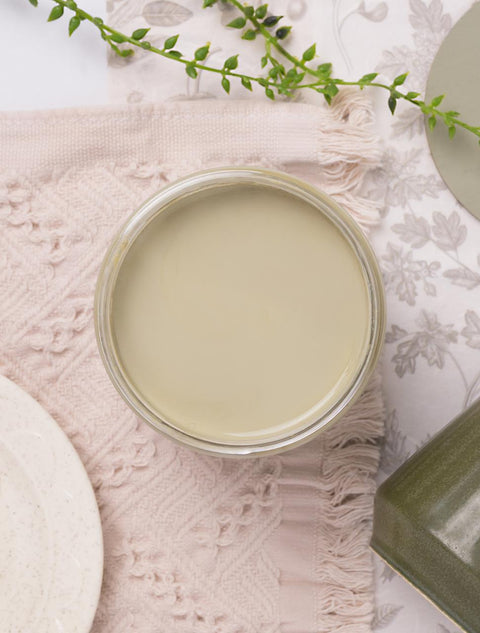 A jar filled with smooth, pale green paint sits on a textured pink cloth, surrounded by greenery and ceramic plates.