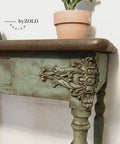 Close-up of a vintage wooden table with ornate floral carvings on its legs and edges. The table is painted in distressed green. A small terracotta pot with a plant sits on the table corner. The words "by ZOLD DESIGN" are visible on the wall.