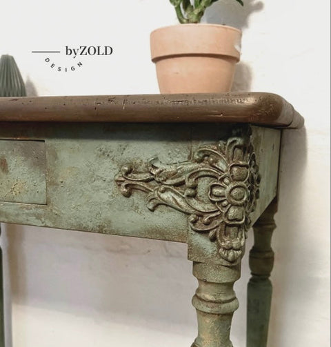 Close-up of a vintage wooden table with ornate floral carvings on its legs and edges. The table is painted in distressed green. A small terracotta pot with a plant sits on the table corner. The words "by ZOLD DESIGN" are visible on the wall.