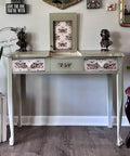 A vintage light green vanity with floral and butterfly-patterned drawers, flanked by two bust sculptures. A circular frame and various decorations are on the wall. A pink chair and potted plants complete the stylish setup.