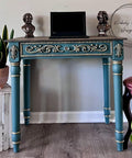 A teal table with ornate gold details stands on a wooden floor. A laptop is placed on top, along with two small bronze busts. A plant is visible to the side.
