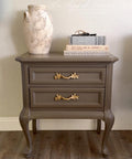 A taupe-colored vintage nightstand with ornate gold handles stands on a wooden floor. On top, there is a textured ceramic vase and two stacked books titled "ART AND LIFE IN AMERICA" and "MICHELANGELO.