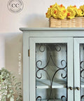 A light blue cabinet with ornate black metal doors stands against a white wall. On top, a wicker basket filled with yellow flowers is displayed. Green leaves cascade from the left side, and a round wall logo is visible.