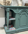 A teal wooden sideboard with ornate carvings on its doors and sides. The interior shelves are lined with colorful floral patterns. A vase with flowers and decorative books rest on top. Text reads "REHAB 2 FAB DESIGNS" on the bottom right corner.