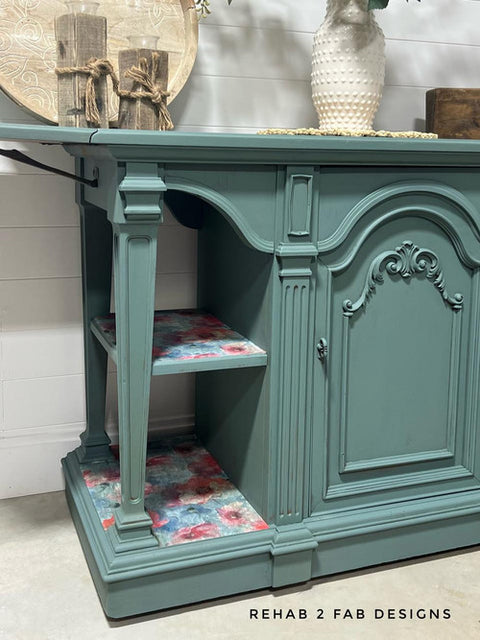A teal wooden sideboard with ornate carvings on its doors and sides. The interior shelves are lined with colorful floral patterns. A vase with flowers and decorative books rest on top. Text reads "REHAB 2 FAB DESIGNS" on the bottom right corner.