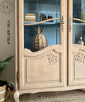 A rustic wooden cabinet with glass doors showcases decorative items, including stacked books, a woven basket, and small ornaments. The cabinet features ornate designs and is placed against a blue-painted interior background. Text reads "Au Naturel.