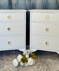 Two white ornate bedside tables with three embossed drawers each, featuring crystal knobs. They stand on a gray fluffy rug with white flowers in between. A textured blue wall is in the background.