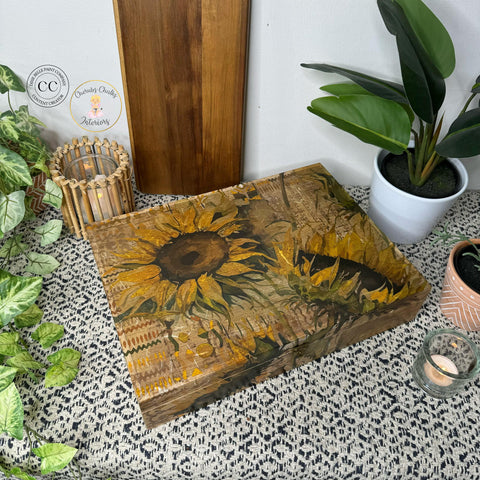 Wooden box with intricate sunflower design, using the Indian Summer Decoupage Paper, rests on a textured surface among potted plants, candles, and rustic decor elements. The scene exudes a cozy, nature-inspired aesthetic with earthy colors and natural materials.