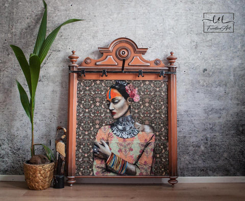 A framed portrait of a woman in traditional attire with colorful makeup, set against a floral background. The frame rests on the floor next to a small plant in a wicker pot and a decorative bird sculpture.