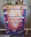 Purple, pink, yellow, and orange painted dresser with white crystal pulls on the top drawer and gold knobs on the remaining 4 drawers. Lani's Celestial Dream Transfer has been applied on all of the 5 drawers, along with an image of a moth and roses. Staged with a rustic picture frame and a gold clock on top. There is a single lit candlestick on the ground.