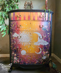 A colorful wooden dresser painted with a celestial theme, featuring a sun, moon, and stars. The dresser has ornate handles, sits in a room with leafy plants, and a single candle rests on top.
