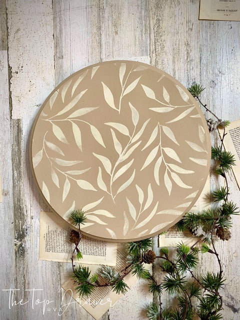 Tan painted lazy susan with the Leaf Me Alone stencil applied in white. Staged with book pages and a garland with small pinecones.
