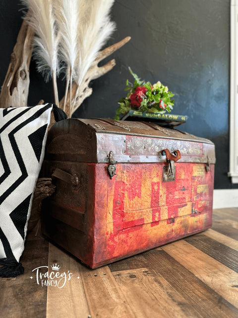 Brown trunk with the Life decoupage paper applied on the front. Staged next to a black and white striped pillow, with a book and flowers on top of the trunk.