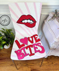 A chair with a striped pattern and a design featuring red lips with a bite mark. The words "LOVE BITES" are written in bold pink and red letters, with a heart shape replacing the "O". There's a small plant and a decorative cabinet nearby.