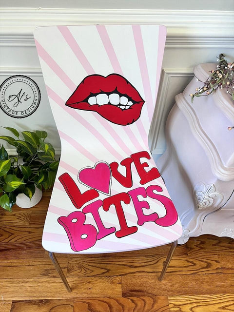 A chair with a striped pattern and a design featuring red lips with a bite mark. The words "LOVE BITES" are written in bold pink and red letters, with a heart shape replacing the "O". There's a small plant and a decorative cabinet nearby.