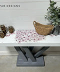 A white and gray wooden table with a crisscross base is decorated with a metal watering can full of green foliage, a woven basket, and three candles. Lilac flowers are on the table's white surface using the Magnificent Magnolia Transfer. The words "REHAB 2 FAB DESIGNS" are on the wall above.