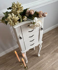 A white, vintage-style jewelry armoire with ornate handles and floral decorations on top, including large ivory and pink blooms. Two lit candles in brass holders sit on the light wood floor nearby.
