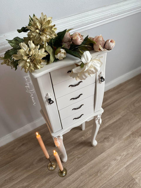 A white, vintage-style jewelry armoire with ornate handles and floral decorations on top, including large ivory and pink blooms. Two lit candles in brass holders sit on the light wood floor nearby.