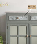 A light green cabinet with intricate patterns on the top drawers and gold knobs. "Magnolia" is labeled with an arrow pointing to the drawer. The cabinet stands on a wooden floor with a potted plant nearby.