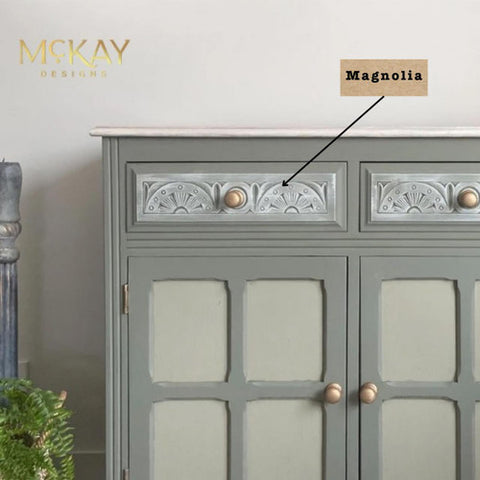 A light green cabinet with intricate patterns on the top drawers and gold knobs. "Magnolia" is labeled with an arrow pointing to the drawer. The cabinet stands on a wooden floor with a potted plant nearby.