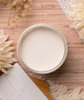 An open jar of cream-colored paint on a wooden surface, surrounded by dried flowers and textured fabric accents. The cream appears smooth and white, with a rustic and natural aesthetic.