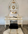 A vintage white vanity with an ornate mirror sits against a wall. The vanity has gold accents and two drawers. A black cushioned stool is placed in front on a white rug. A bed with plush pillows is reflected in the mirror.