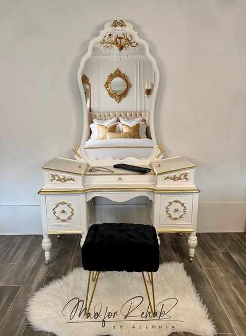 A vintage white vanity with an ornate mirror sits against a wall. The vanity has gold accents and two drawers. A black cushioned stool is placed in front on a white rug. A bed with plush pillows is reflected in the mirror.