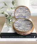 A decorative open box with intricate blue and white patterns is displayed on top of a stack of books. A small glass jar with green leaves is placed beside the box. The books are titled "Beautiful Australian Homes," and "Graphic Simplicity."