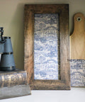 A still life featuring a pair of old binoculars on top of a book, next to a framed textile art piece and a wooden cutting board with the Ming Vase Decoupage Paper applied, displayed on a shelf.