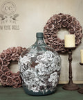 A large, intricately decorated ceramic floral vase is centered against a light wall. It is flanked by two metal candlesticks with candles and adorned with two wreaths made of dusty rose-colored flowers. The backdrop features a logo for "Low Flying Angels."