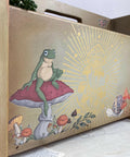 Wooden magazine holder with Cottagecore Mushrooms Silkscreen Stencil and Mr. Toad's Happy Place Transfer applied on the front of one of the sides. Staged with a faux string of pearls hanging off the side.