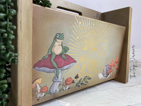 Wooden magazine holder with Cottagecore Mushrooms Silkscreen Stencil and Mr. Toad's Happy Place Transfer applied on the front of one of the sides. Staged with a faux string of pearls hanging off the side.