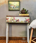 White chalk painted table with a dark wood top. Mr. Toad's Transfer has been applied on the front of the table. Staged with a book and a metal watering can, with a pothos inside, on top.